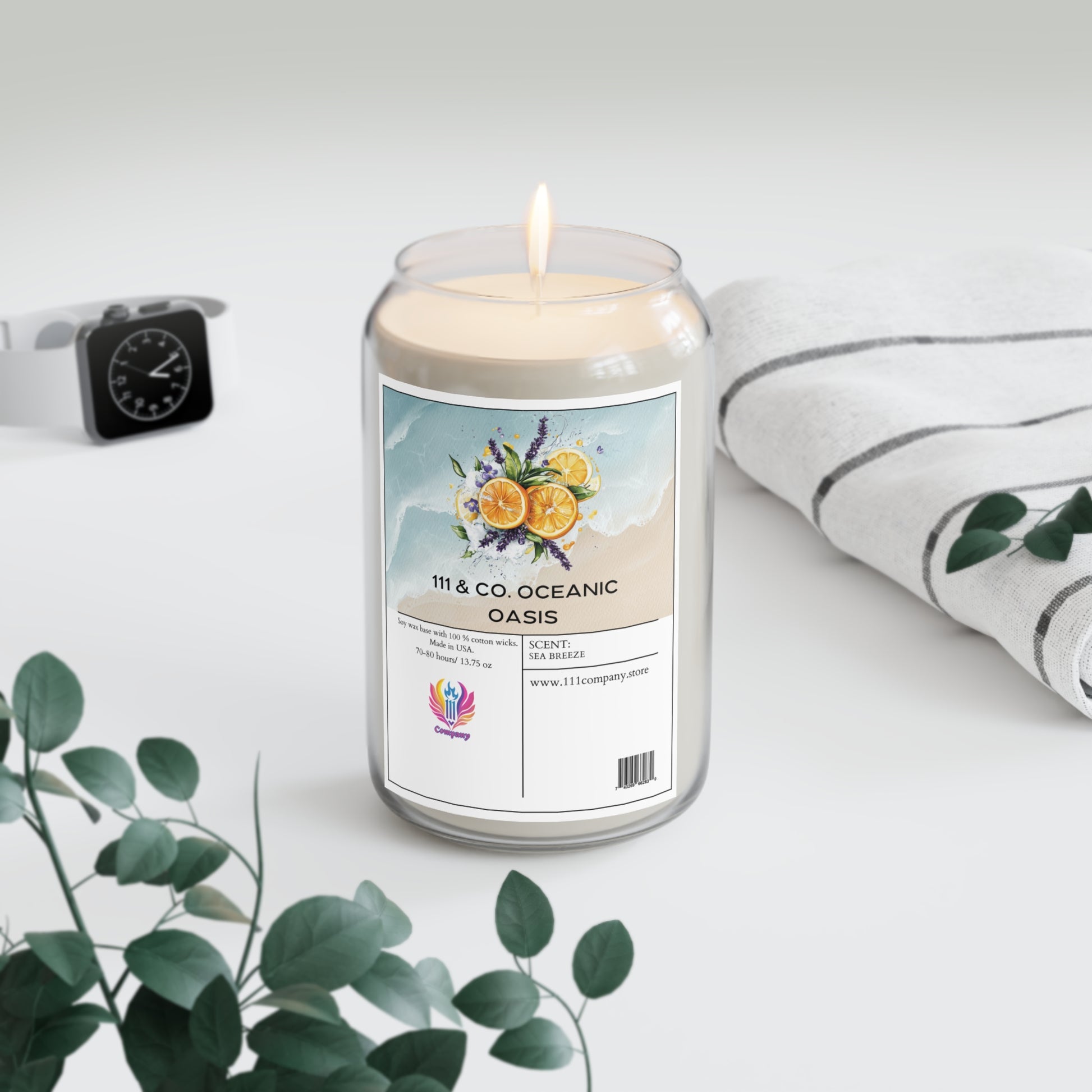 A 111 & Company Oceanic Oasis Sea Breeze soy wax candle from 111 & Company, with citrus, lavender, and cedarwood notes, sits in a glass jar on a light surface. Nearby are green eucalyptus leaves, an Apple Watch, and a striped white cloth, creating a calm and fresh ambiance.