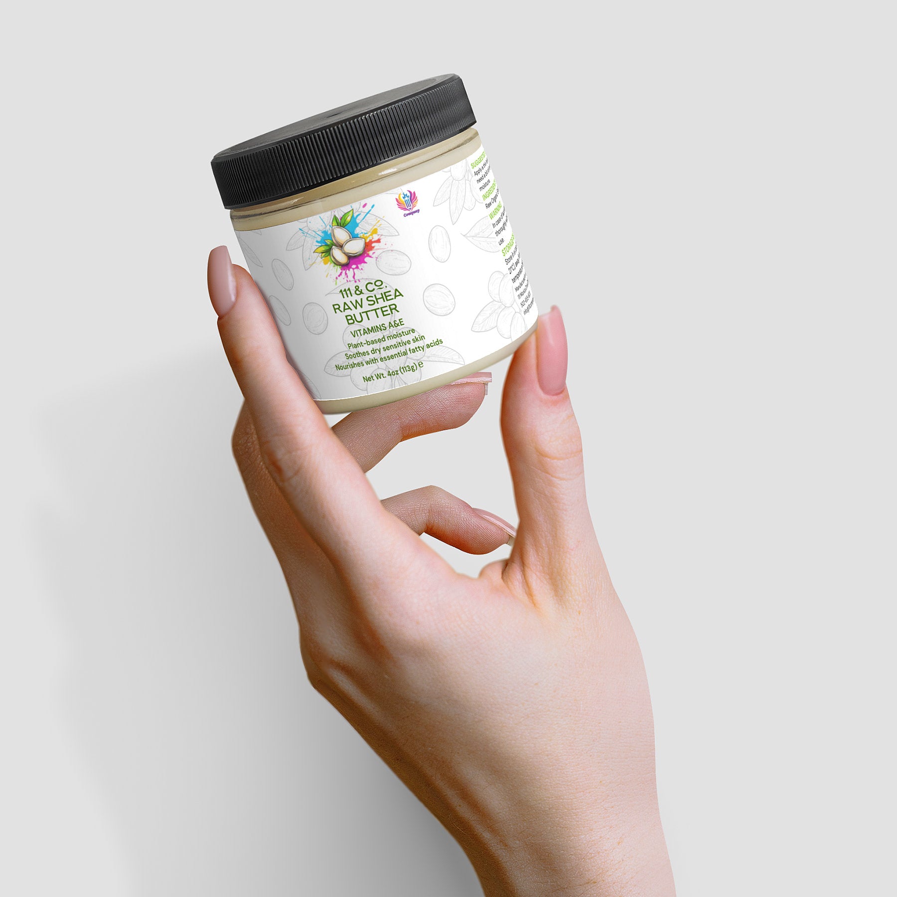 A pale hand holds a small jar of 111 & COMPANY RAW SHEA BUTTER by 111 & Company against a light gray background. The jar, topped with a black lid, flaunts a vibrant label with flowers and leaves. Manicured nails add elegance to this plant-based moisturizer display.