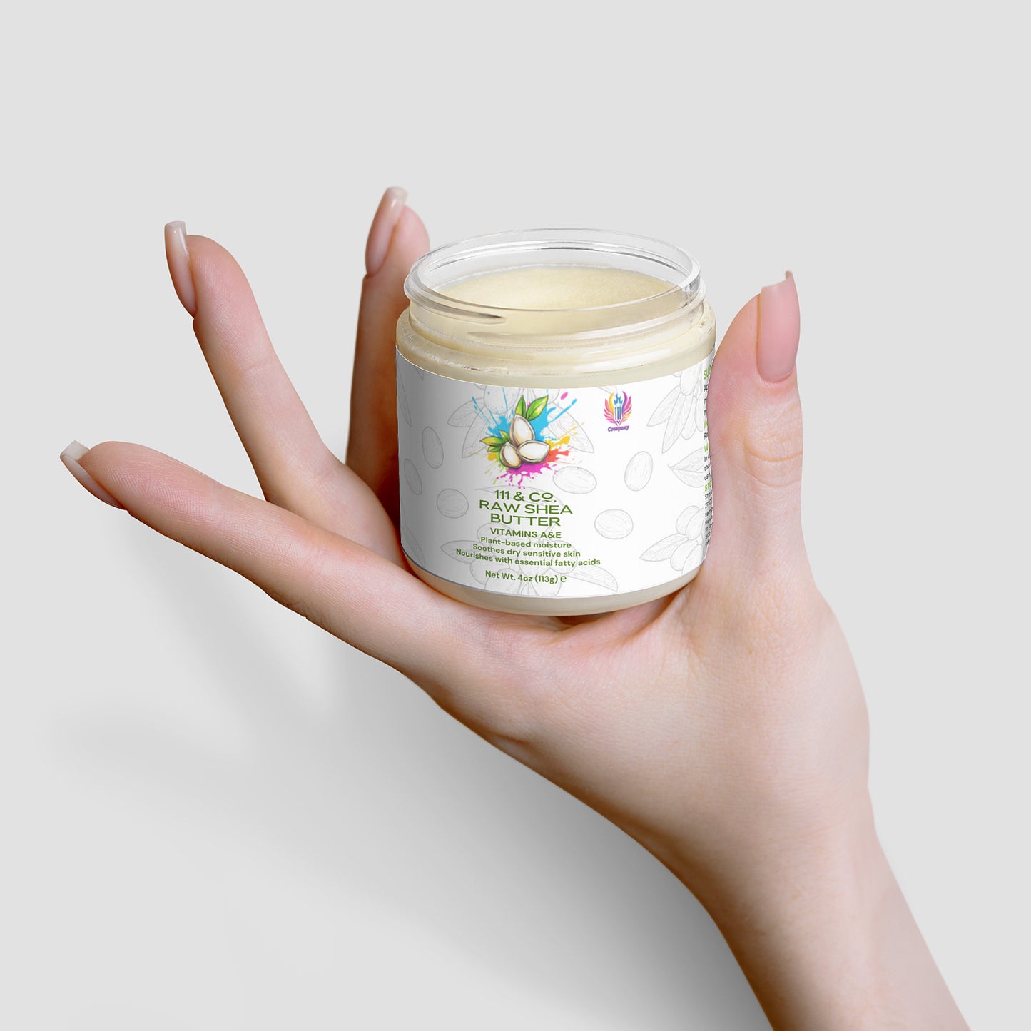 A hand with long nails holds a clear jar of  111 & COMPANY RAW SHEA BUTTER. The label, showcasing colorful fruit graphics and text, emphasizes this plant-based moisturizer. Set against a soft gray backdrop, the creamy texture inside enhances its minimalist appeal.