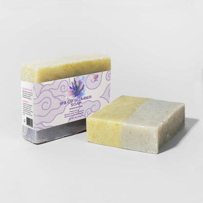 Two handcrafted soap bars are shown, one wrapped in a label reading 111 & Company Slumber Soap by 111 & Company, with lavender and fir needle imagery. The unwrapped bar reveals a marbled yellow and gray-green texture. Both rest on light gray surfaces, casting gentle shadows.