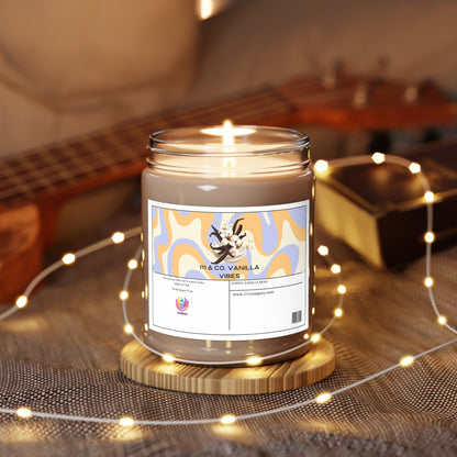 A 9oz vanilla bean-scented soy wax candle from 111 & Company, labeled 111 & COMPANY VANILLA VIBES, sits in a clear jar with abstract orange and blue designs on a wooden coaster. Warm fairy lights create a cozy ambiance, and blur your vision of the guitar that leans against the background wall.