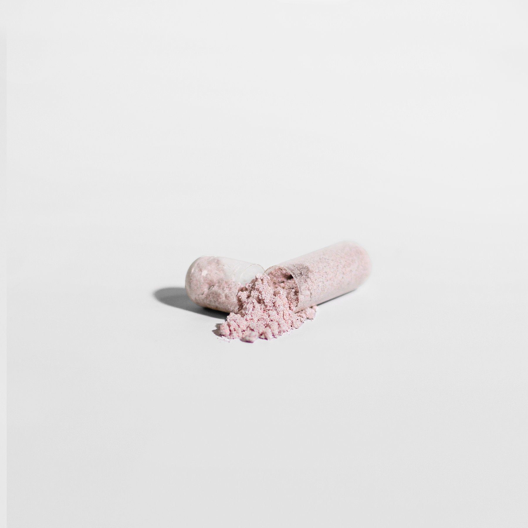 A close-up of an 111 & COMPANY VISION SUPPORT CAPSULE by 111 & Company on a white background, showing light pink powder spilling out. The cylindrical capsule, split into two halves, lies horizontally with the powder forming a small pile and casting a soft shadow.