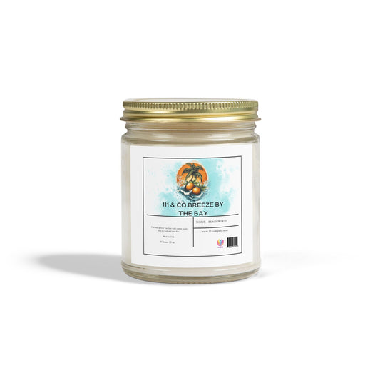 The Printify 111 & Co: Breeze by the Bay Beachwood Scented Candle features palm trees and water artwork on a clear glass jar with a gold lid. Made from eco-friendly coconut apricot wax, this 9oz candle promises a coastal escape while casting a soft shadow.