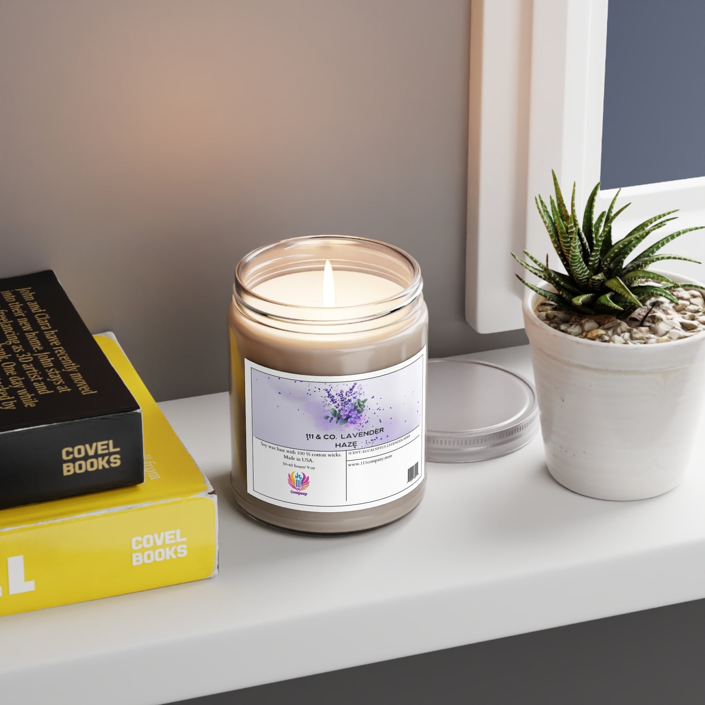 A lit Printify 111 & Company Lavender Haze Eucalyptus Lavender Herb Candle made from a soy wax blend sits in a glass jar on a white shelf. Next to it is a small green succulent in a white pot and books with yellow and black covers. The candle label shows lavender illustrations, while above, a white-framed picture peeks into view.