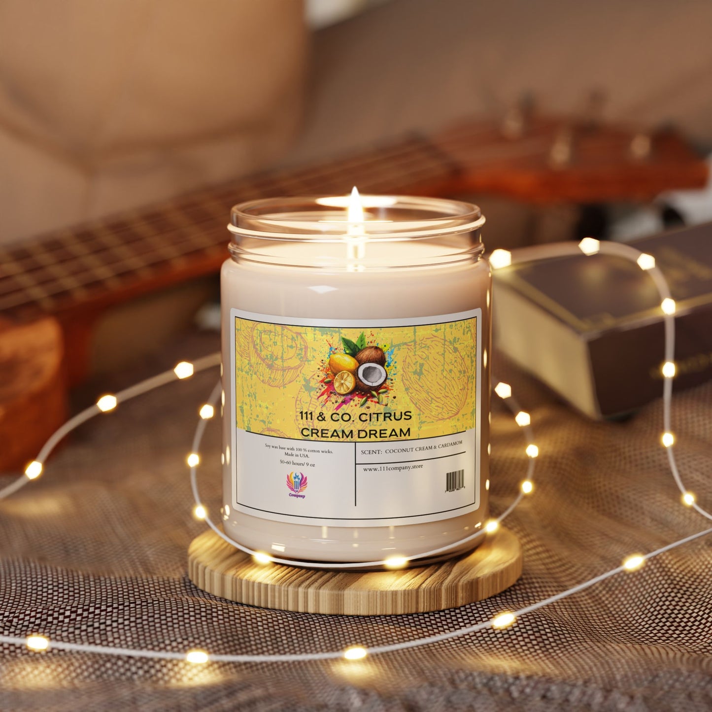 A lit Printify candle, labeled 111 & Company Citrus Cream Dream Coconut & Cardamom Scented Candle, rests on a wooden base amid delicate fairy lights. A blurred ukulele, an open book, and soft fabric in the background create a cozy ambiance reminiscent of a tropical paradise.