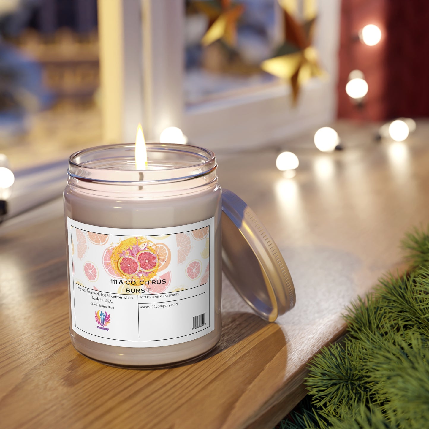 A 111 & COMPANY CITRUS BURST soy wax candle with a pink grapefruit scent from Printify sits in a jar on wood. Its label shows citrus slices. A window features star decorations and blurred string lights, creating a cozy feel. Greenery is visible to the right.