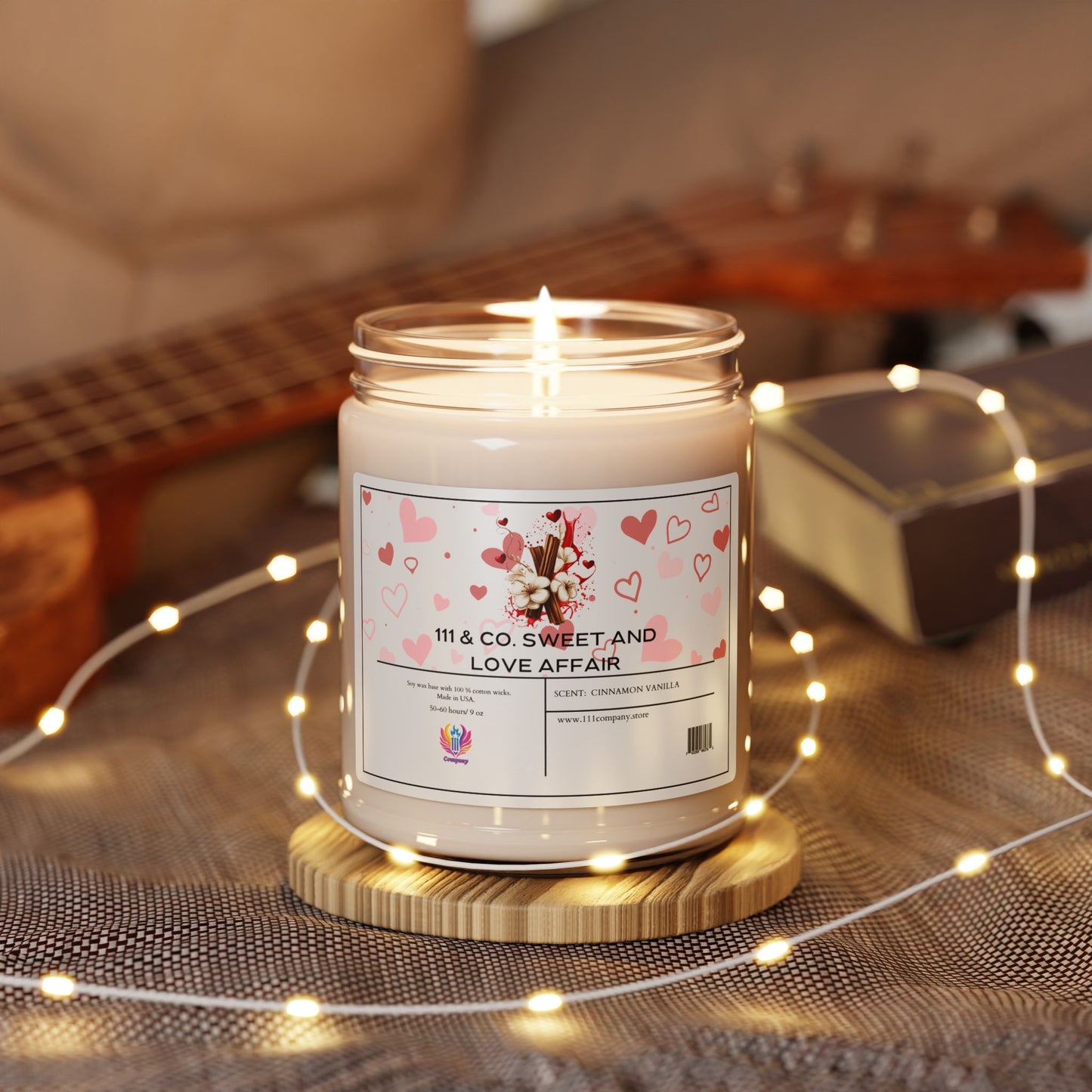 A 9oz Printify 111 & Company candle named Sweet and Love Affair, featuring a pink heart design, rests on wood, exuding a warm cinnamon vanilla aroma. Fairy lights glow nearby, with a blurred ukulele and closed book in the backdrop.