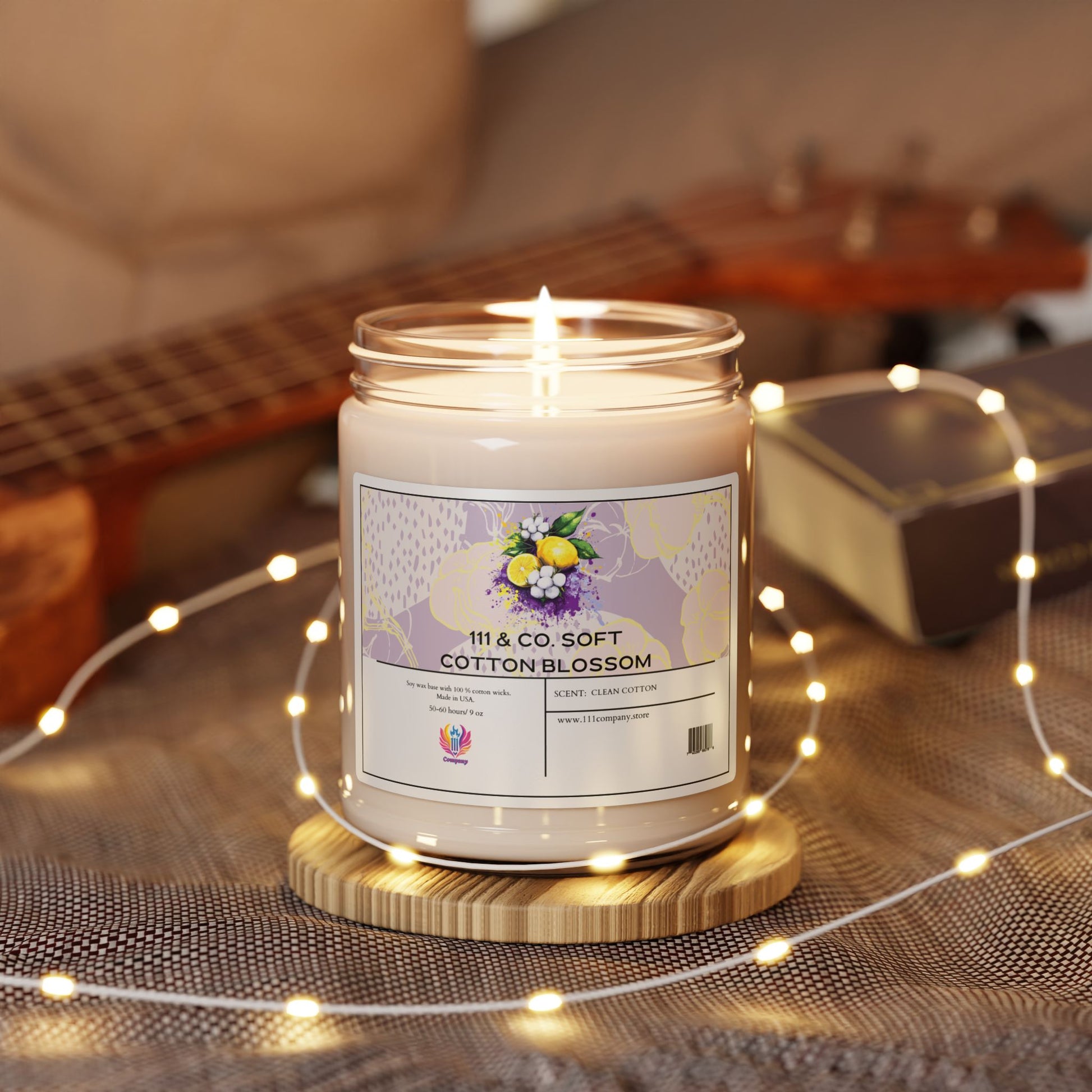 A floral-decorated 111 & Company Soft Cotton Blossom Scented Candle (9oz soy wax) by Printify is lit on a wooden coaster, surrounded by white string lights and a ukulele backdrop, creating a cozy ambiance with its long burn time.