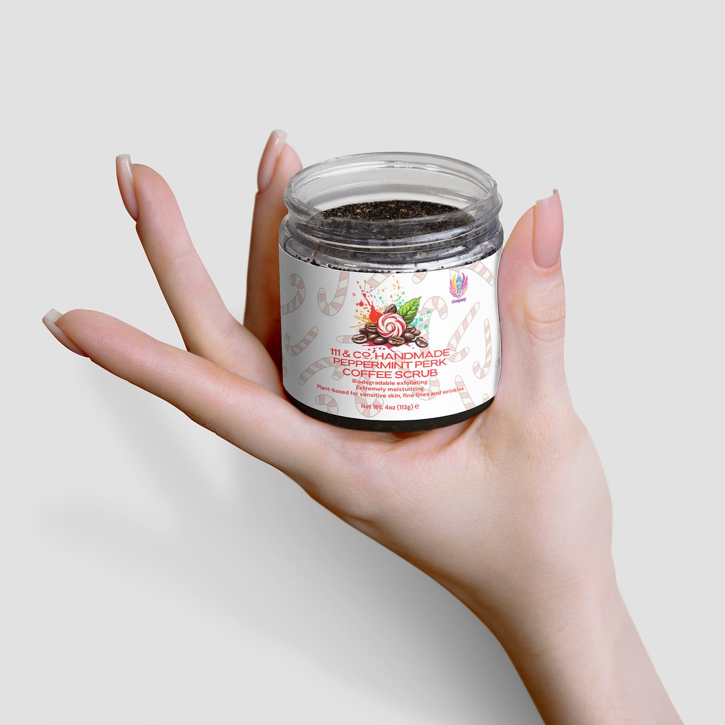 A hand with long, manicured nails holds a transparent jar of 111 & Companys Handmade Peppermint Perk Coffee Scrub against a white background. The jar has a colorful label featuring a peppermint candy design, and the dark exfoliating scrub is visible inside.