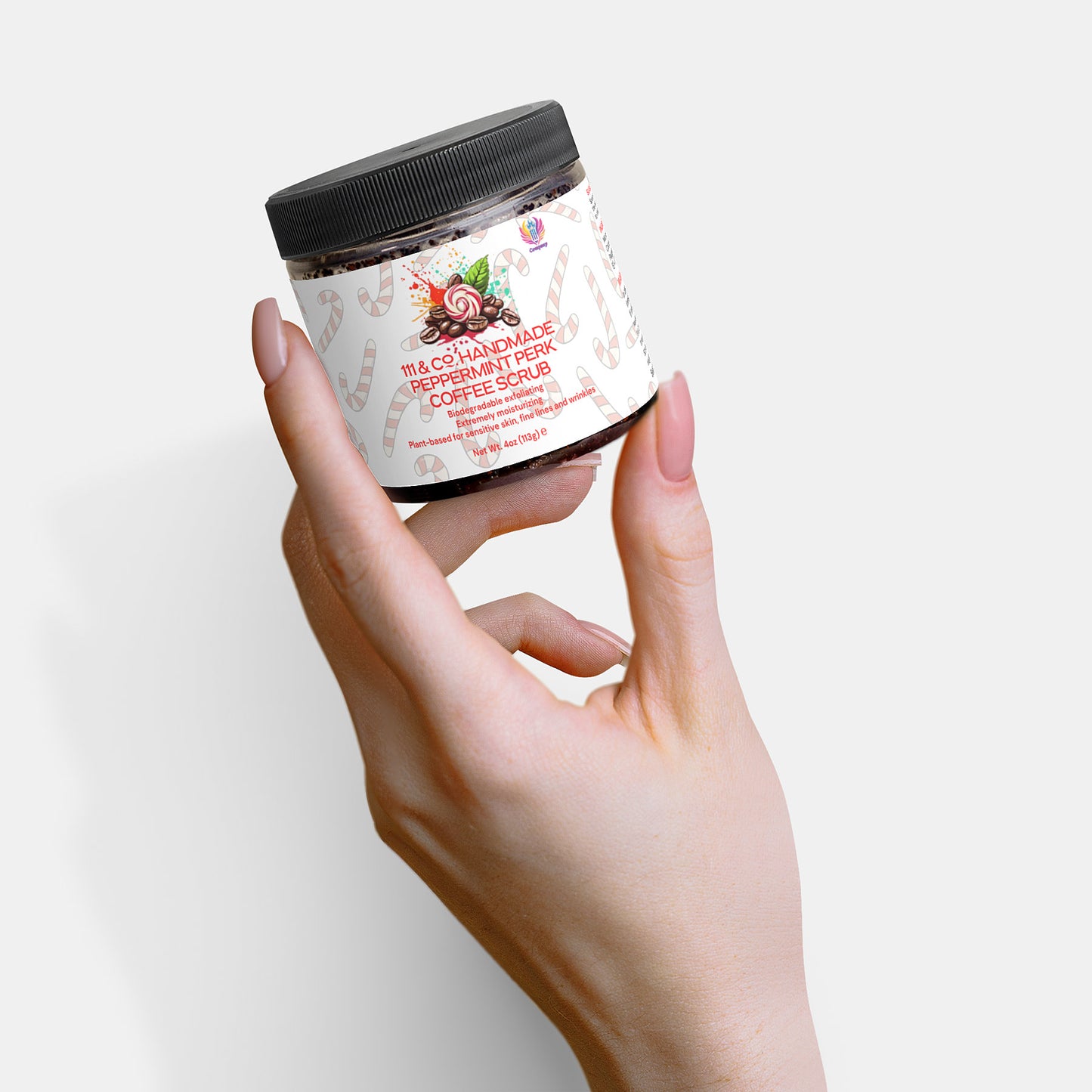 A light-skinned hand holds a jar labeled 111 & COMPANY HANDMADE PEPPERMINT PERK COFFEE SCRUB. The jar has a black lid, white label with peppermint and coffee bean imagery, and is designed for exfoliation. A plain white background highlights the hand and antioxidant-rich scrub.