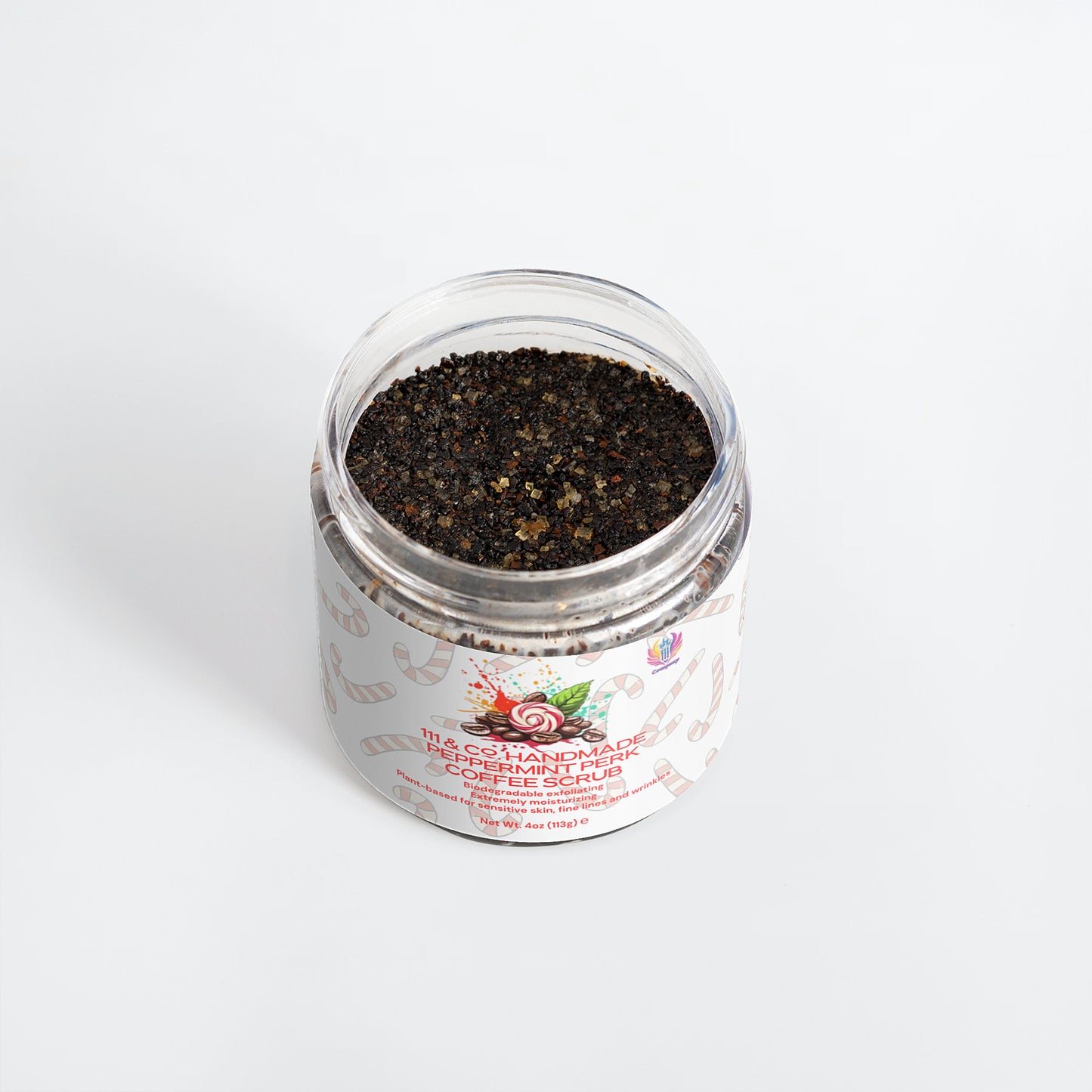 A clear jar with a white lid holds a coarse, dark mix labeled 111 & Company Peppermint Perk Coffee Scrub. The label features candy canes and vibrant text. This antioxidant-rich scrub offers a peppermint aroma, adding an aromatic contrast against its plain white background.