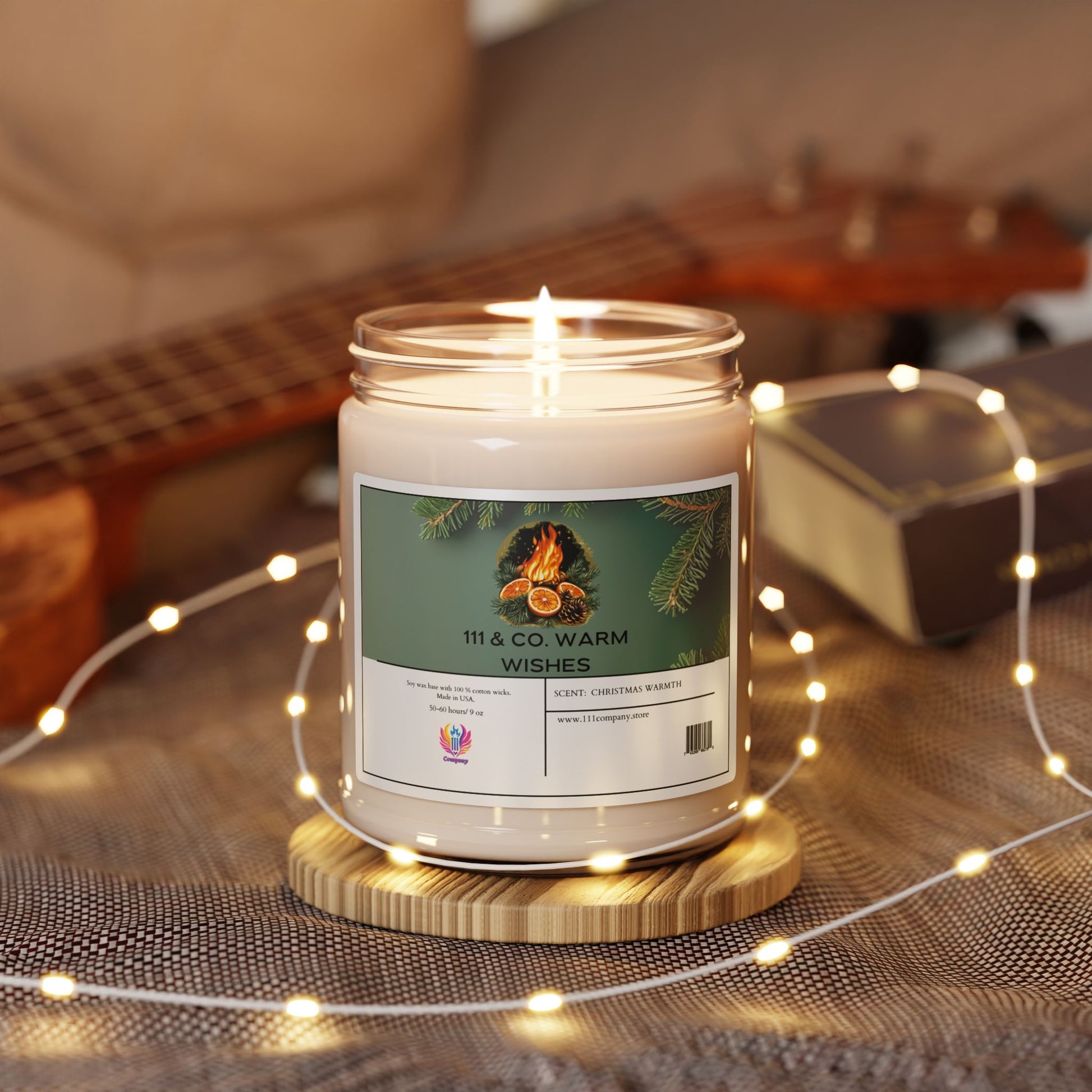 The Printify 111 & COMPANY WARM WISHES CHRISTMAS WARMTH scented candle rests on a wooden coaster. A ukulele and book sit on soft mesh fabric with warm fairy lights draped around, filling the air with a cozy holiday fragrance.