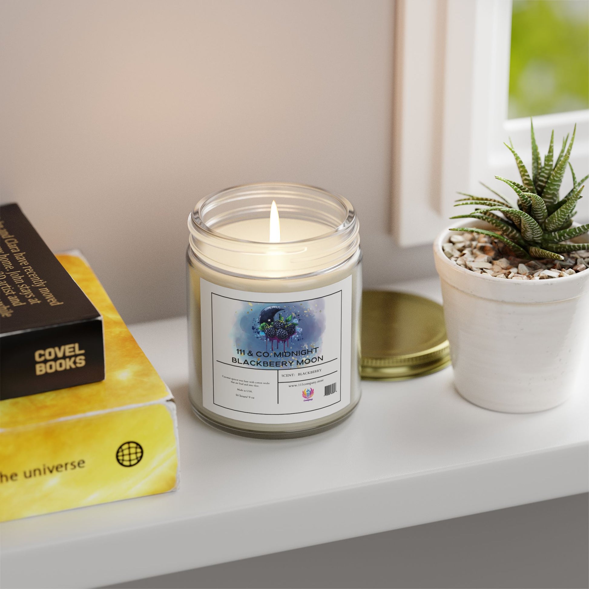 A lit 111 & COMPANY MIDNIGHT BLACKBERRY MOON Scented Candle in a glass jar sits on a white shelf beside a potted succulent. The candle, with Coconut Apricot Wax, is next to books like THE UNIVERSE and a window revealing greenery outside. The brand is Printify.