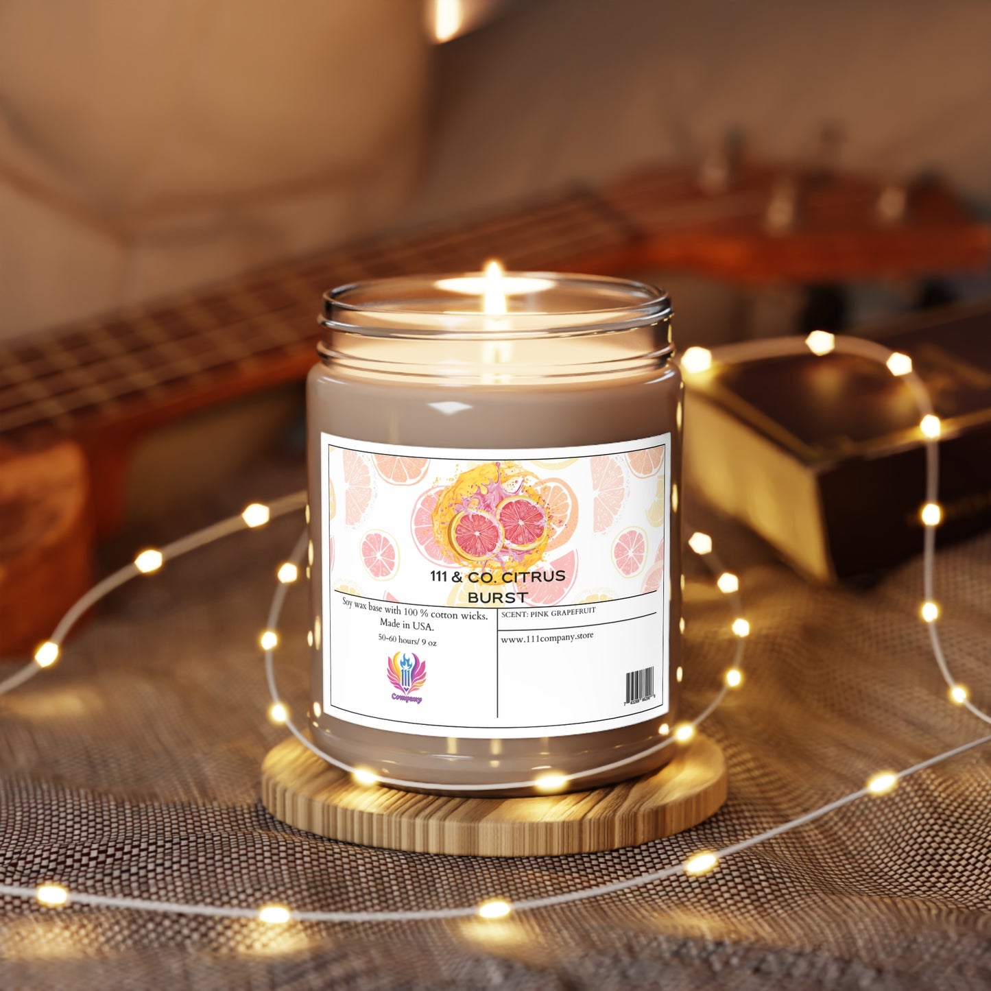 An inviting ambiance is created by a lit 111 & Company Citrus Burst Pink Grapefruit soy candle in a glass jar, resting on textured cloth. The Printify label displays vibrant grapefruit images with warm fairy lights and the blurred fretboard of a guitar as the backdrop.