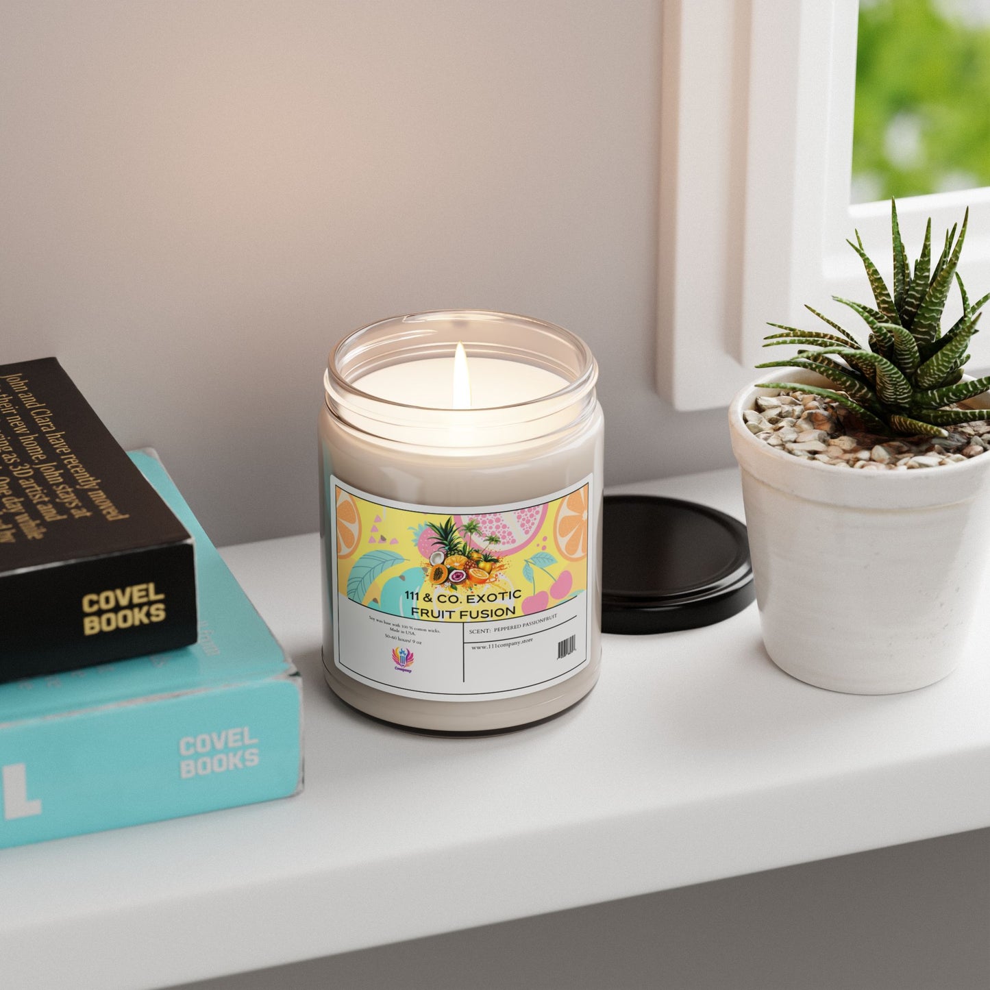 A lit Printify 111 & Company Exotic Fruit Fusion scented candle in a glass jar rests on a white windowsill. Its label suggests an exotic aroma. Nearby are an open black lid, a small potted succulent, and books with Covel Books partially visible on their spines.