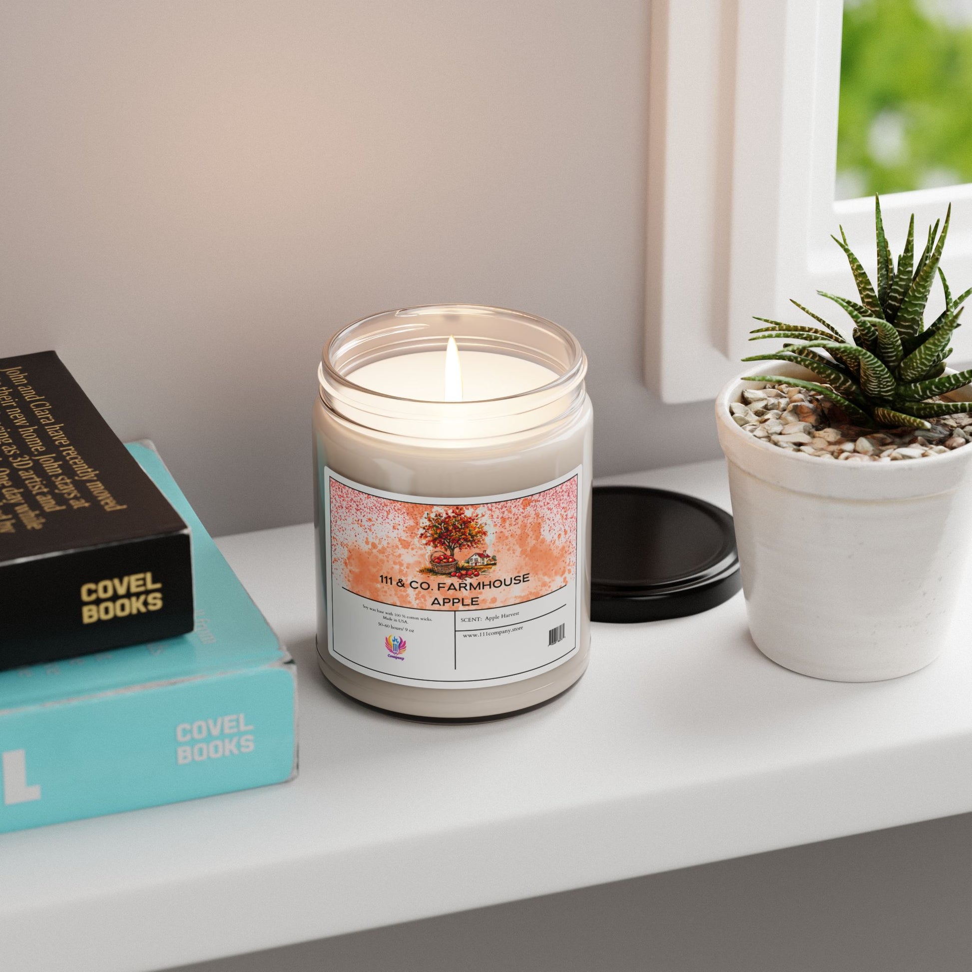 A lit 111 & Company Farmhouse Apple Harvest soy candle by Printify sits on a white shelf with two books and a small potted succulent. The eco-friendly candle is in a glass jar with a black lid nearby, as soft daylight filters through the window, creating a cozy autumn ambiance.