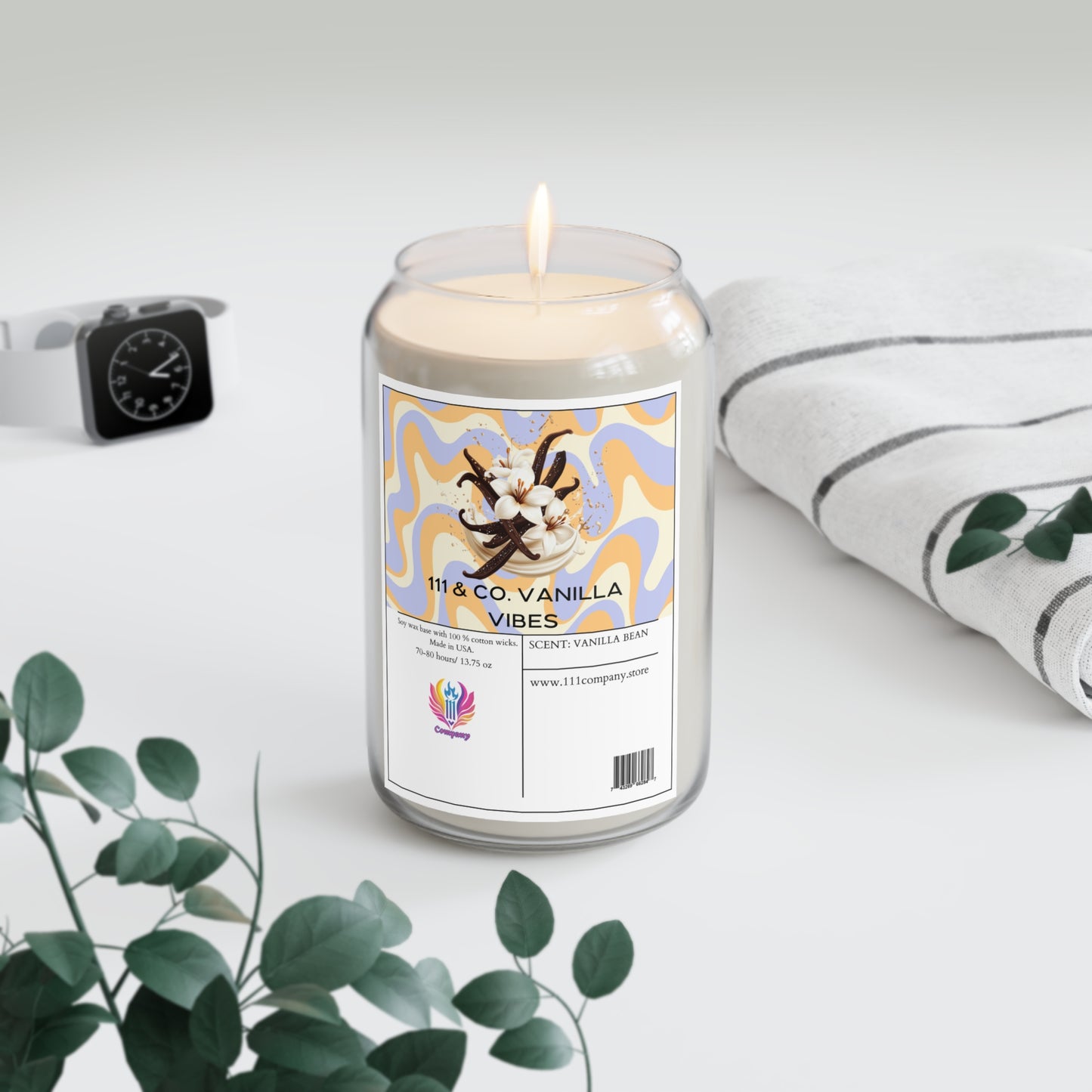 A 111 & COMPANY Vanilla Vibes candle, resembling a soda can and enriched with vanilla and tonka bean scents, sits with a striped towel, smartwatch, and eucalyptus branches on a soft white surface.