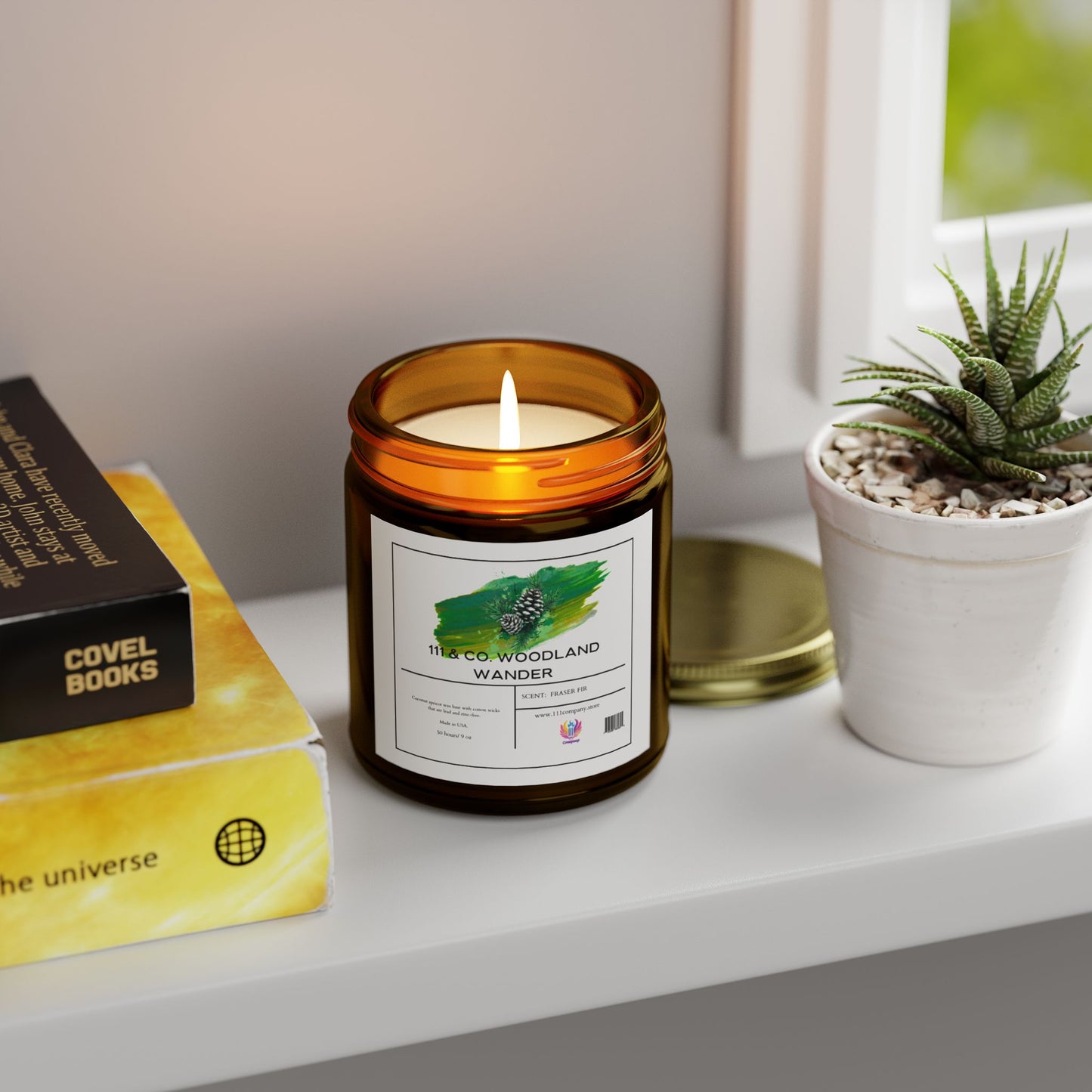 A lit 111 & COMPANY Woodland Wander Fraser Fir candle from Printify, made of Coconut Apricot Wax and housed in a brown jar, sits on a white shelf beside a potted succulent. Nearby are two books—one black, one yellow—under soft natural light from a closed window.