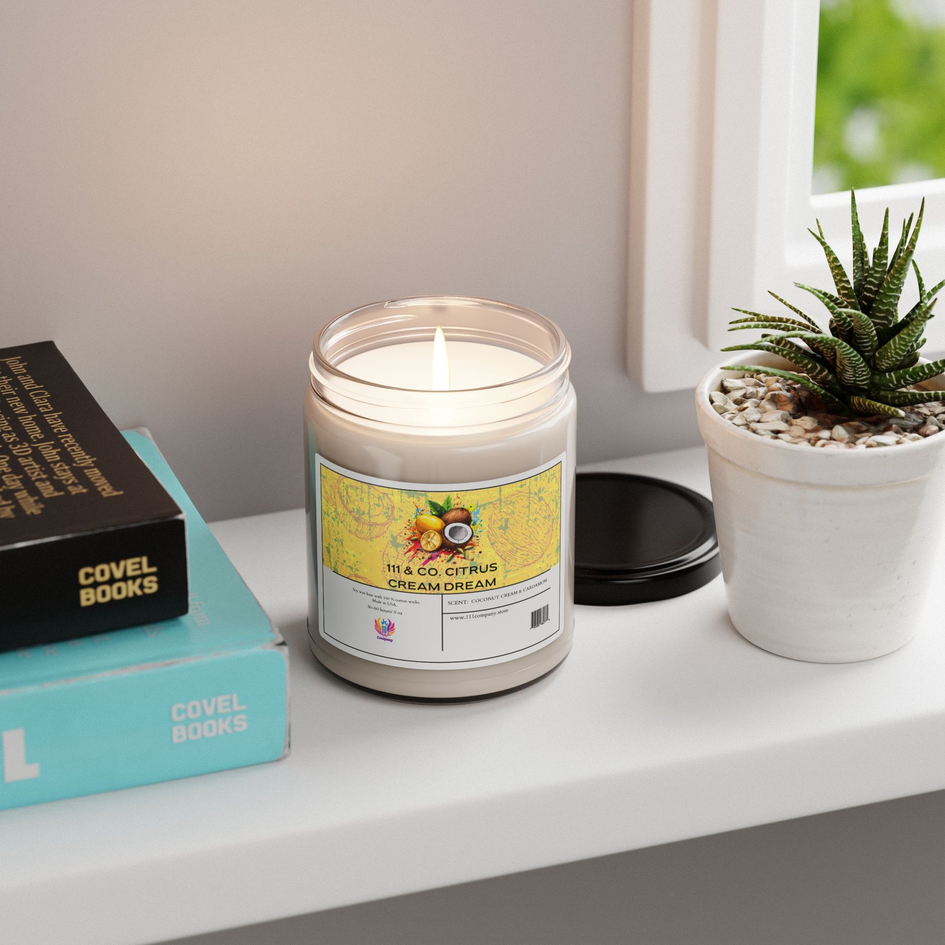 The 111 & COMPANY Citrus Cream Dream candle by Printify, a soy wax blend with coconut and cardamom scent, sits on a white shelf next to a potted succulent. Nearby, a book from Covel Books is stacked as natural light enhances the tropical fragrance. The candles lid lies flat nearby.