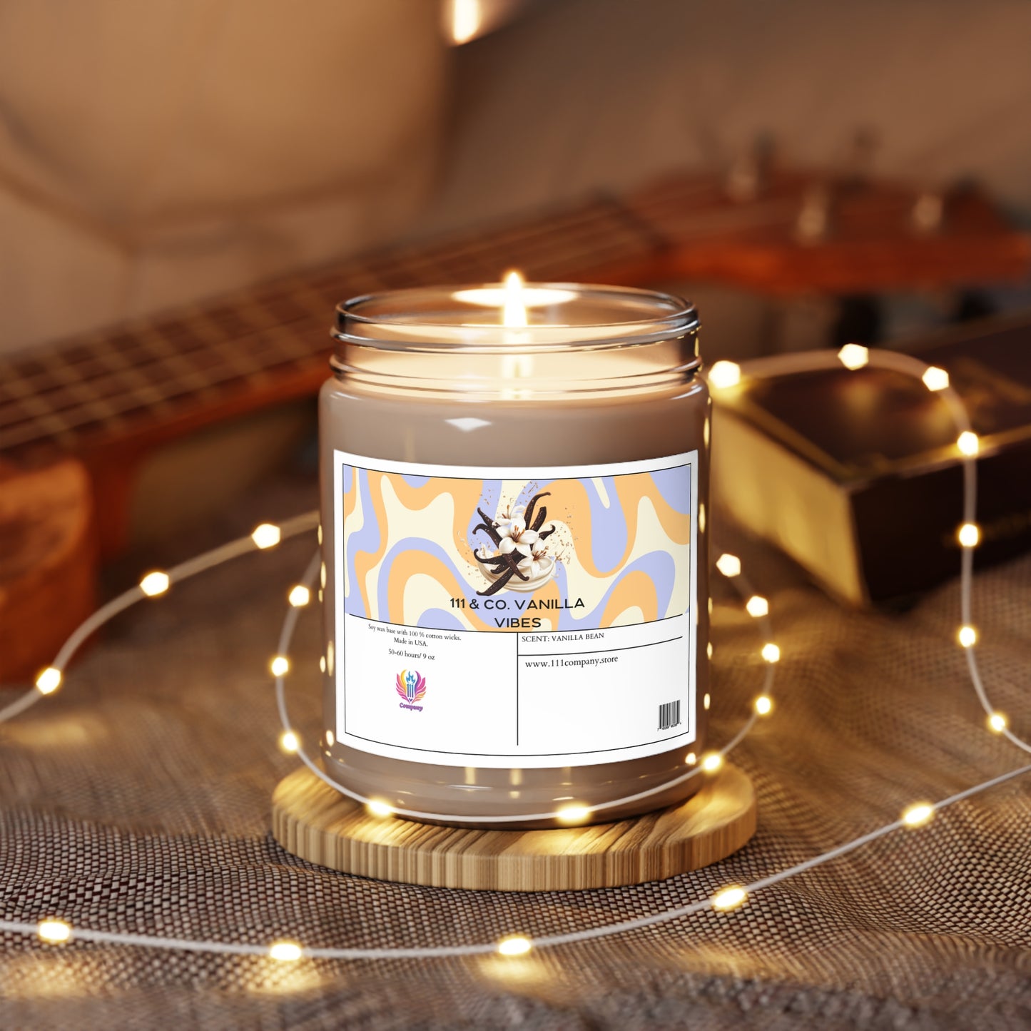 A 9oz vanilla bean-scented soy wax candle from Printify, labeled 111 & COMPANY VANILLA VIBES, sits in a clear jar with abstract orange and blue designs on a wooden coaster. Warm fairy lights create a cozy ambiance, and blur your vision of the guitar that leans against the background wall.